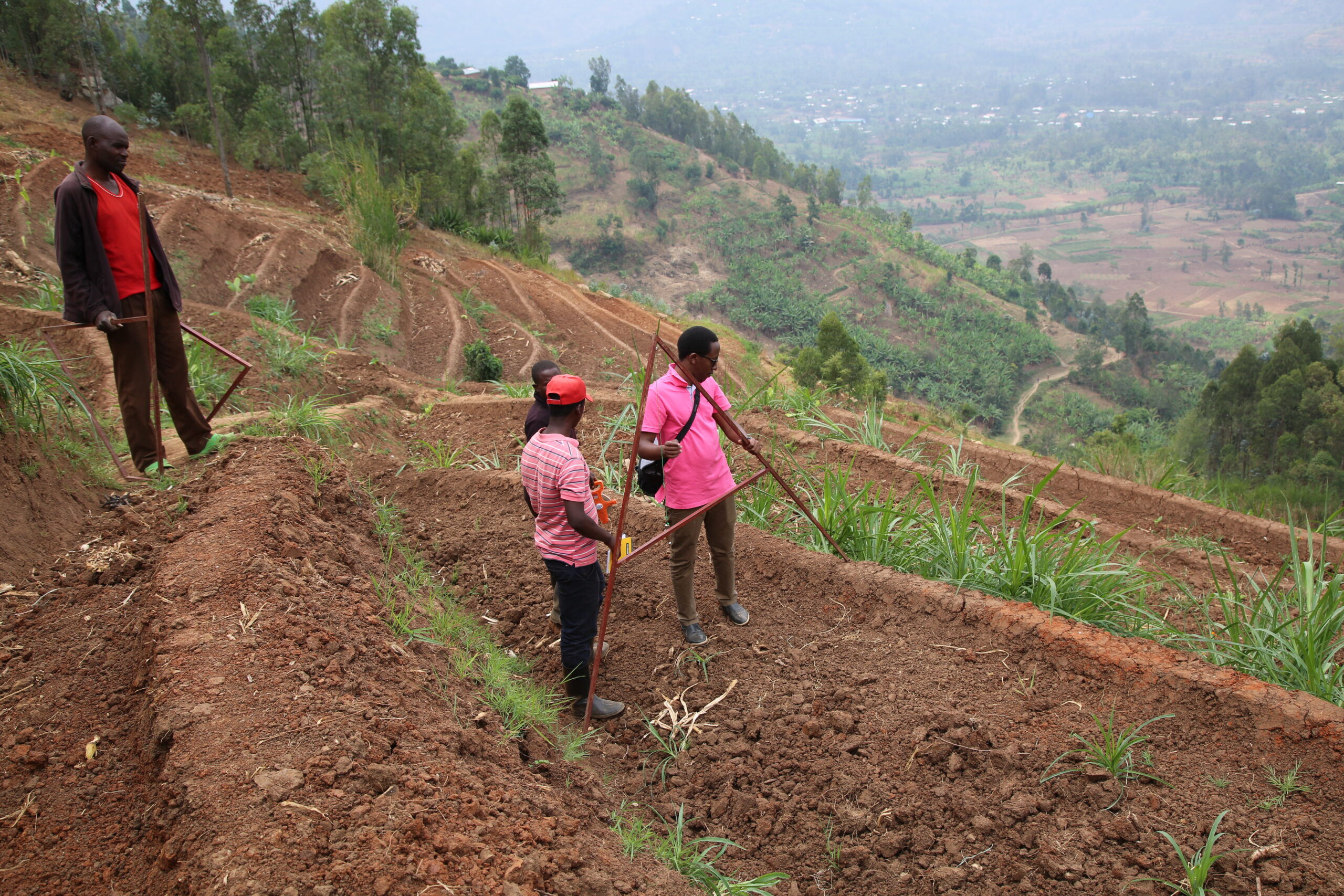 A landscape photograph taken on a hillside in Rwanda. There are four people in the foreground of the image, who are surveying the land. The ground is mostly soil with a few shrubs of grass growing. There are trees in the background.