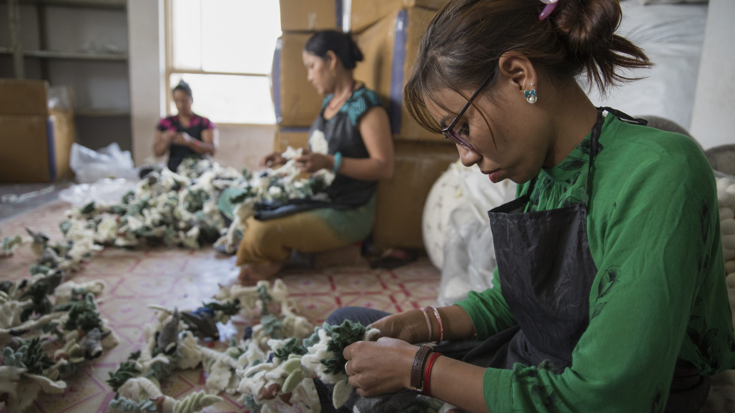 Une photo de trois femmes assises sur le sol, les jambes croisées. Elles cousent des objets d'artisanat. À l'arrière-plan, des boîtes sont empilées.