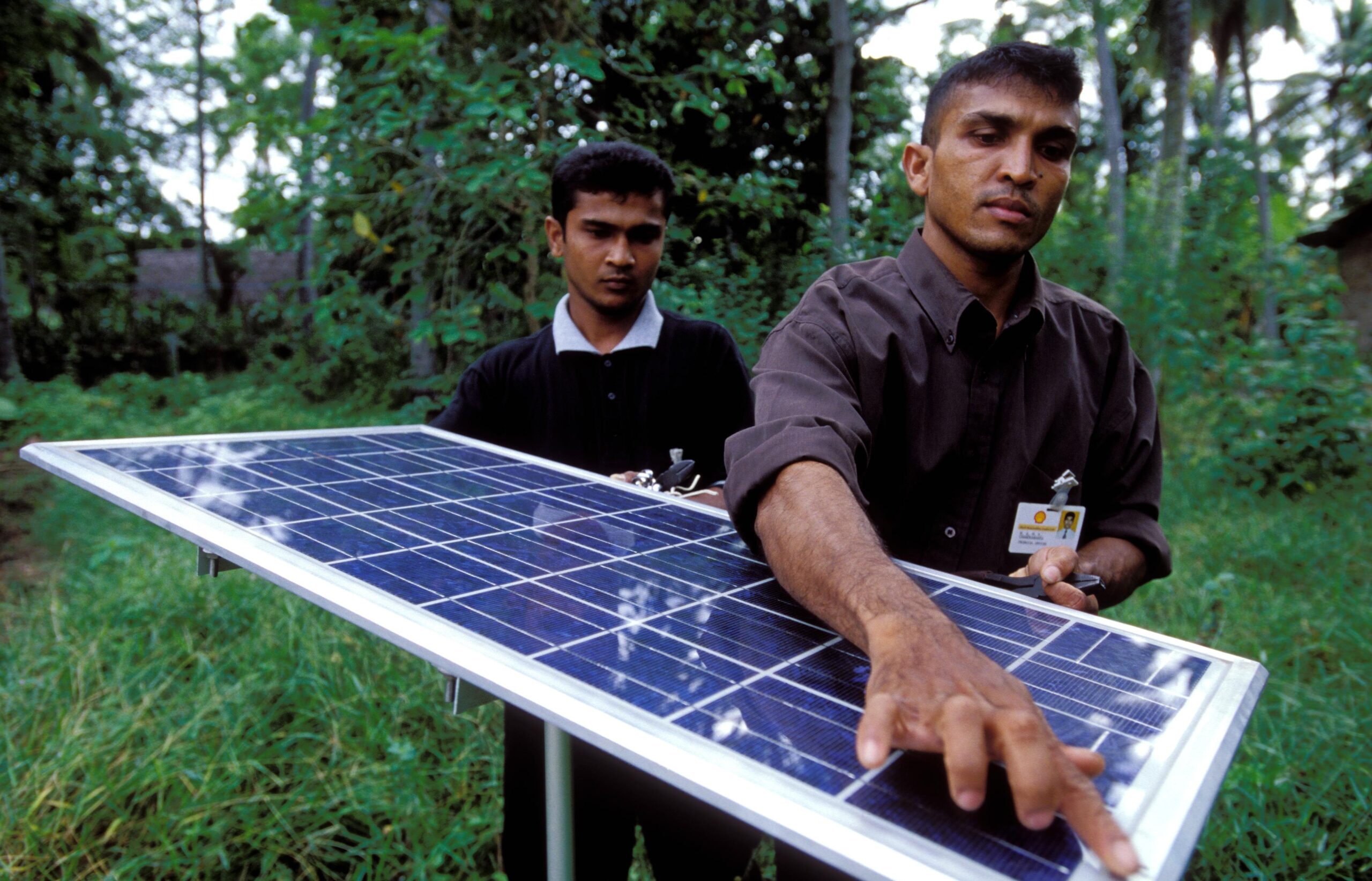 A photo of two men looking at a solar panel. One is pointing to the corner. There are green trees in the background.