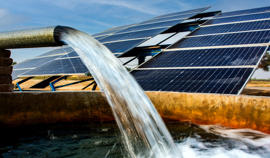 A photo of a solar panel. A pipe is in front of it and is gushing water into a tank. The ground in the background looks dry.