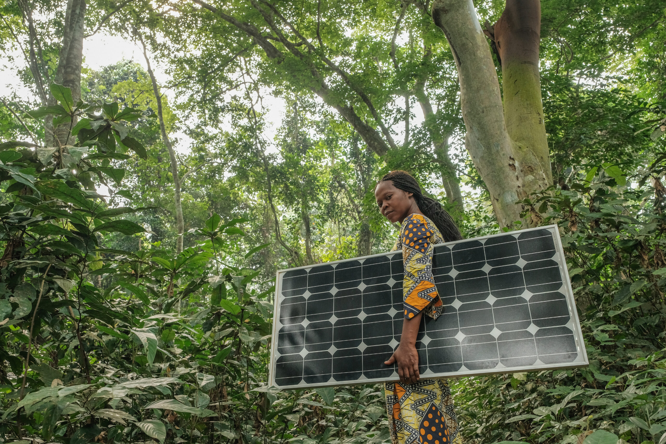 A photo of a woman carrying a solar panel in a wooded area. The sky is lined with trees and the woman looks towards the camera.