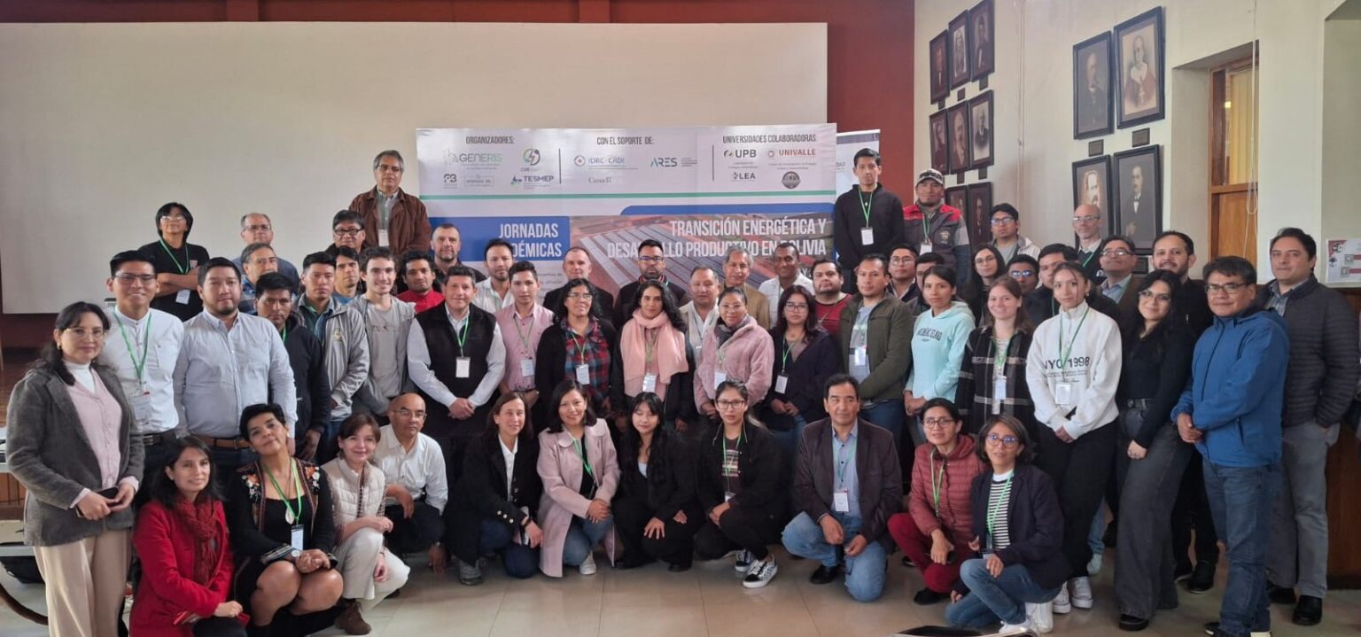 Group photo of approximately 50 conference participants standing and kneeling in front of a large banner that reads “Transición Energética y Desarrollo Productivo en Bolivia.” The participants include men and women of diverse ages, wearing conference badges and casual business attire. The banner behind them displays logos of the GENERIS Project, UMSS, IDRC–Canada, UPB, UNIVALLE, and other partner institutions. The setting appears to be an auditorium or lecture hall with framed portraits on the walls.
