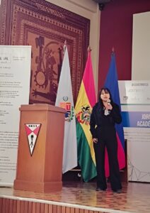 A woman dressed in black speaks into a microphone while standing on a stage next to a wooden podium with the UMSS logo. Behind her are three flags: Bolivia, Cochabamba department, and another blue and red flag. Academic and sponsor banners are visible in the background. 