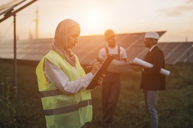 A woman wearing a hijab and high-visibility vest writes on a clipboard at a solar energy site, with two male workers in hard hats discussing plans in the background during sunset.