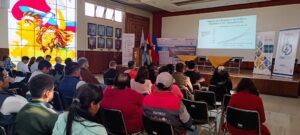 Audience members attend a presentation in a well-lit auditorium with a large stained-glass window and portraits on the wall. A speaker is presenting a slide on the influence of distance on SME investment in R&D. The stage features national and institutional flags, and banners for the GENERIS and CUIE projects are visible. The room is filled with attendees seated and engaged with the talk.