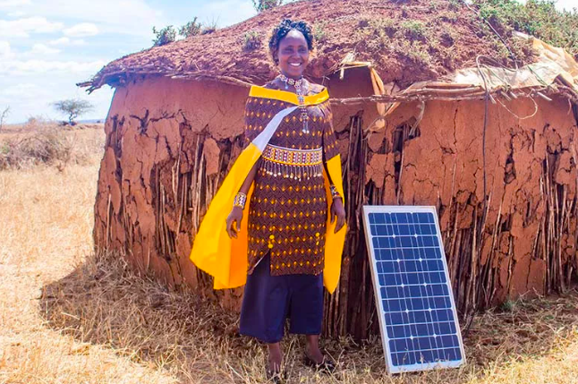 Smiling woman in traditional attire stands next to a small solar panel outside her earthen home in a dry rural landscape.