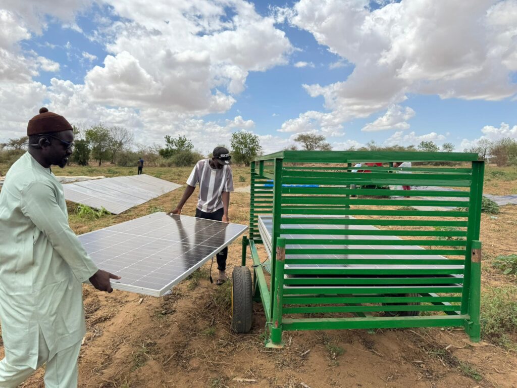 A photo of two men lifting a solar panel and moving it onto a crate. They are in a farmed field, and the sky is blue with clouds. 