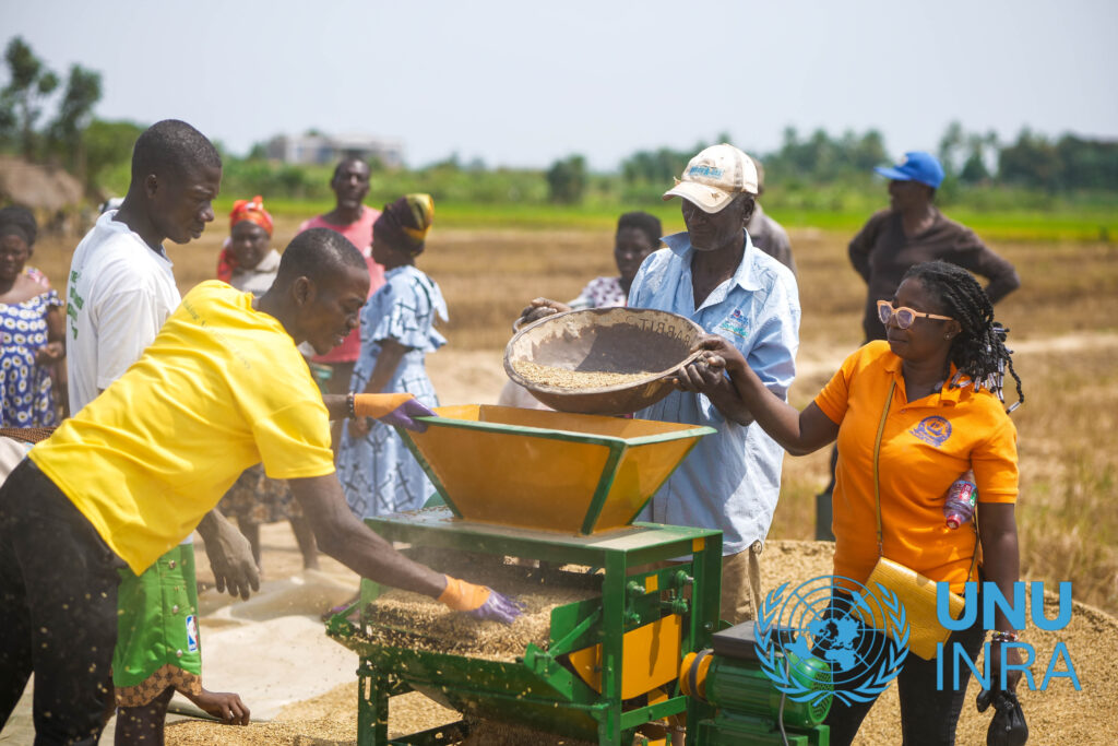 A photo of a group of people standing around a new machine which has been developed. One person holds a bowl with grains and tips it into the machine for processing, and another person stands at the other side, collecting the milled grains. 