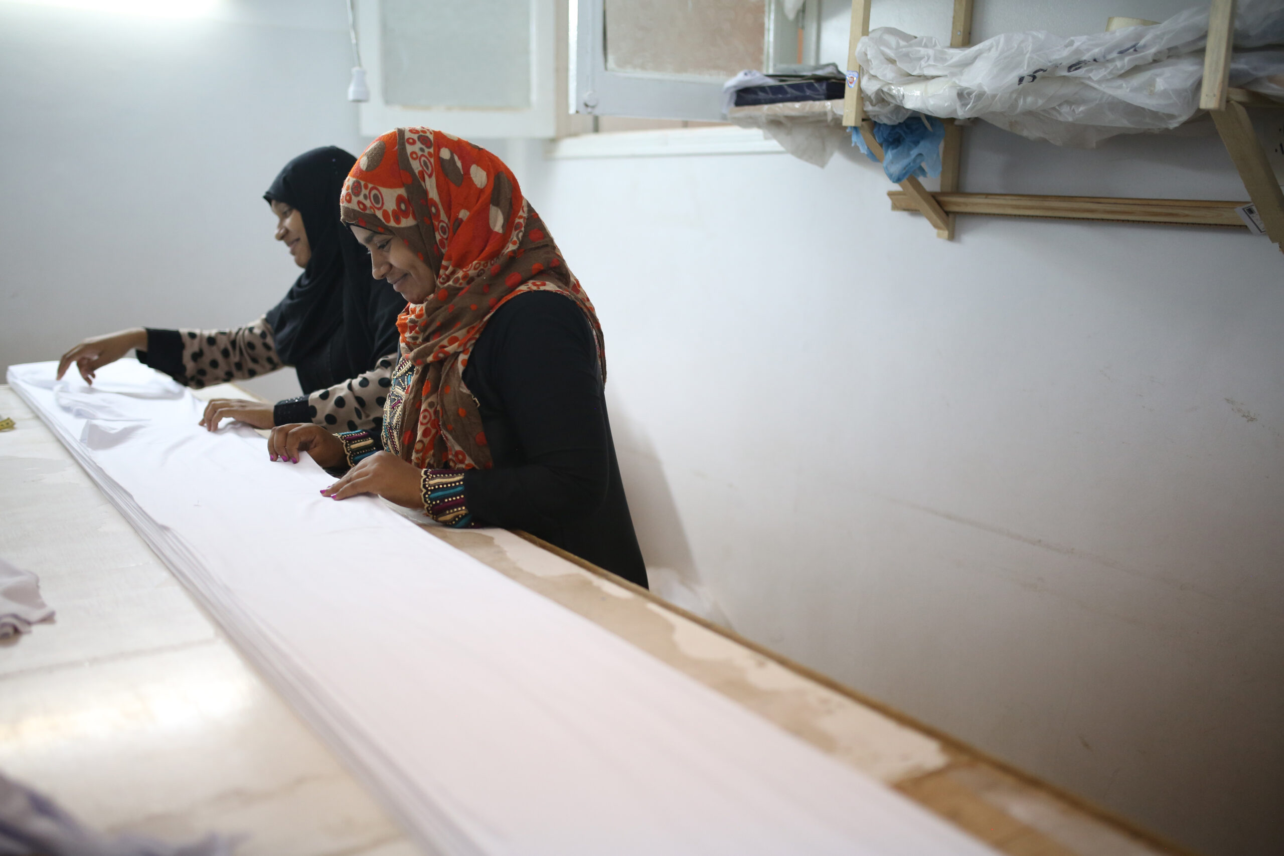 Two women are standing by a table in a factory, folding a long piece of white fabric.