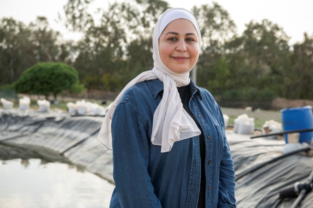 A photo of a woman smiling at the camera, standing outside a reservoir. In the background there are green trees.